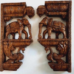 Puede incluir: Un par de soportes de pared rectangulares de madera con detalles tallados a mano. Cada soporte muestra una figura central de elefante sobre una plataforma, con un motivo de pavo real tallado en la base y molduras decorativas en la parte superior.