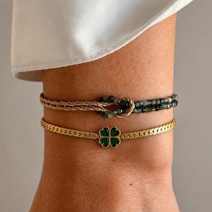 Könnte beinhalten: Zwei zusammen getragene Armbänder. Das obere Armband besteht aus einer goldfarbenen gedrehten Kette, die mit einem Strang kleiner, dunkelgrüner facettierter Perlen verknotet ist. Das untere Armband ist eine goldfarbene Fischgrätenkette.