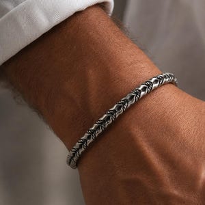 Op de afbeelding: Een zilverkleurige metalen armband met een doorlopend, herhalend blad- of veerachtig patroon met geoxideerde donkere details in de verdiepte delen. De band heeft een afgerond, getextureerd oppervlak dat in een strak, uniform ontwerp om de pols valt.