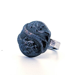 May include: A silver-toned ring featuring a detailed, dark-colored dragon design. The dragon is intricately carved on a round, slightly textured surface. The ring band is a simple, polished silver.