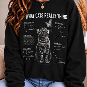 Puede incluir: Una sudadera negra de cuello redondo con el texto blanco WHAT CATS REALLY THINK, un gráfico de un gato atigrado mirando a una mariposa y varias ecuaciones físicas que incluyen dinámica de fluidos, fuerza de arrastre y Bernoulli.