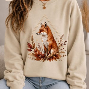 Könnte beinhalten: Ein sandfarbenes Sweatshirt mit Rundhalsausschnitt und einem Grafikdruck eines Fuchses, der inmitten von Herbstblättern und Laub sitzt. Das Motiv ist von einer dünnen, goldfarbenen Rautenlinie umrahmt. Der Text FARBE: SAND steht oben links.