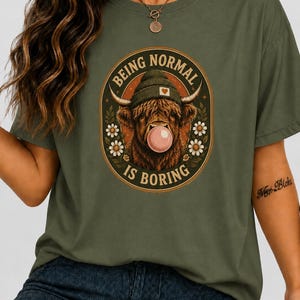 Puede incluir: Una camiseta verde militar con un gráfico de una vaca Highland que lleva un gorro y hace una burbuja rosa. El diseño ovalado incluye margaritas blancas y el texto "BEING NORMAL IS BORING" en una fuente arqueada alrededor de la vaca.