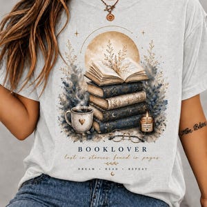Könnte beinhalten: Ein aschefarbenes T-Shirt mit einer Grafik aus einem Bücherstapel, einer dampfenden Tasse mit Herz, einer Kerze und einer Brille unter einem Mond. Der Text lautet: BOOKLOVER, lost in stories, found in pages, und DREAM READ REPEAT mit einem Halbmond.
