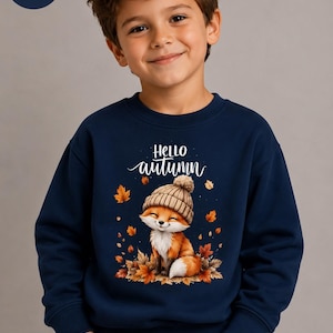 Könnte beinhalten: Ein dunkelblaues Langarm-Sweatshirt mit einem Grafikdruck eines Fuchses, der eine beige Strickmütze trägt. Der Fuchs ist von orangefarbenen und braunen Herbstblättern umgeben. Der Text "Hello autumn" ist in weißer Schreibschrift aufgedruckt.
