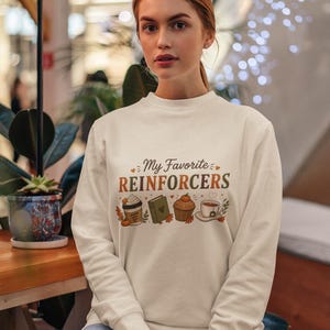 Peut inclure: Un sweat-shirt à col rond de couleur crème avec le texte My Favorite REINFORCERS en lettres marron et orange, accompagné d'illustrations graphiques d'une tasse de café, d'un livre vert, d'un muffin et d'une tasse de thé avec des feuilles d'automne.