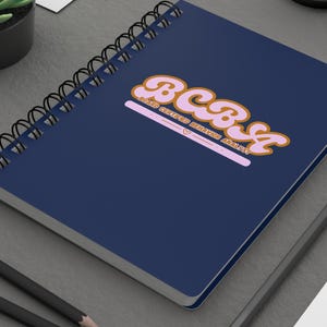 May include: A dark blue spiral-bound notebook featuring the pink and orange text BCBA and Board Certified Behavior Analyst with a small heart icon. The notebook is positioned on a dark gray surface alongside two black pencils and a small succulent.
