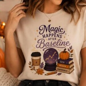 Puede incluir: Una camiseta de color crema con el texto "Magic HAPPENS AFTER Baseline" en morado y marrón. El gráfico incluye una taza de café, una bola de cristal con un gráfico de líneas, una calabaza, una pila de tres libros etiquetados "DATA", "BEHAVIOR" y "REINFORCEMENT", y un lápiz.