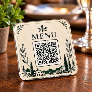 May include: A square, clear acrylic sign featuring a cream-colored background with the word MENU printed in black. The sign displays a central QR code, surrounded by dark green illustrations of olive branches, cypress trees, and a small village.
