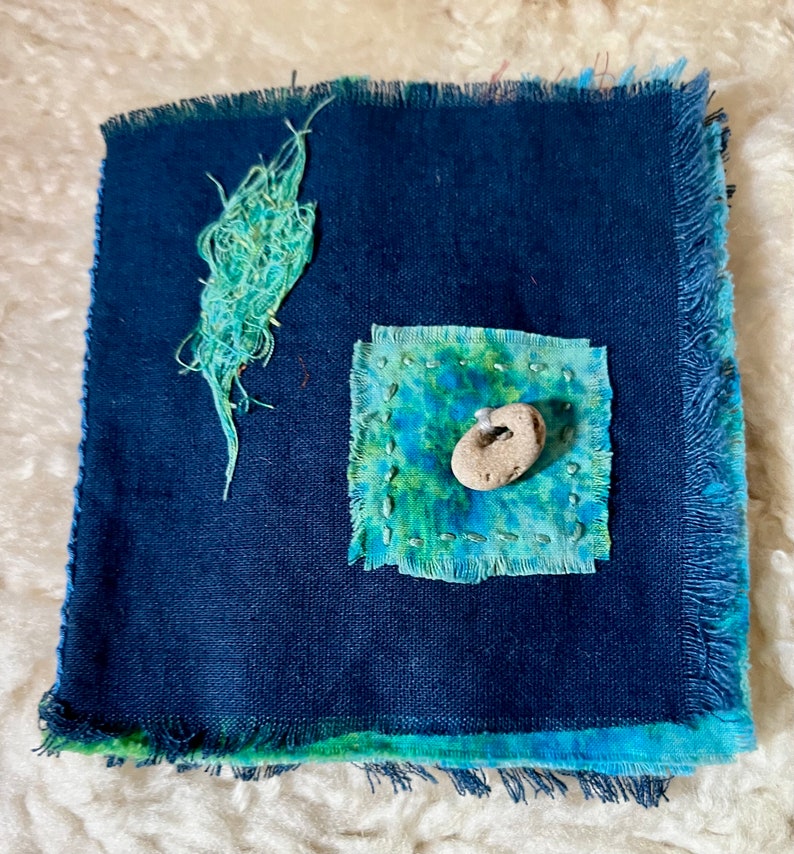Puede incluir: Un peque&ntilde;o diario de tela azul con un borde deshilachado. La portada tiene un parche cuadrado de tela azul y verde con una peque&ntilde;a piedra blanca en el centro. Hay un parche m&aacute;s peque&ntilde;o de tela azul y verde en la esquina superior izquierda.