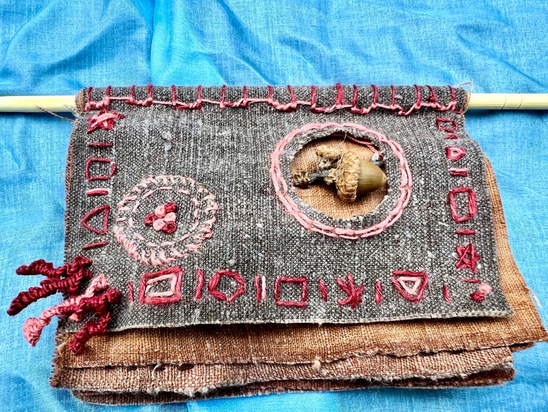 Puede incluir: Un tapiz de pared de tela marr&oacute;n con bordados rosas. El bordado incluye un c&iacute;rculo con una bellota en el centro, un c&iacute;rculo m&aacute;s peque&ntilde;o con un dise&ntilde;o floral y una fila de peque&ntilde;os cuadrados y tri&aacute;ngulos.
