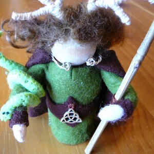Puede incluir: Una muñeca de fieltro hecha a mano con cabello castaño, que lleva una túnica verde, una capa marrón y un collar de plata. La muñeca sostiene una serpiente de fieltro verde y un bastón de madera.