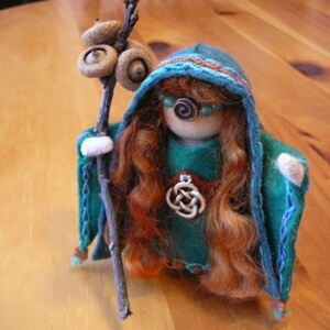 Puede incluir: Una muñeca hecha a mano con cabello largo y rojo, vestida con una capa con capucha azul y verde y sosteniendo un palo con bellotas. La muñeca lleva un vestido verde y un colgante con un nudo celta.