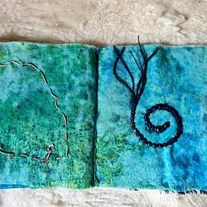 Puede incluir: Un diario de tela hecho a mano con una cubierta de m&aacute;rmol azul y verde. El diario tiene un c&iacute;rculo cosido en la p&aacute;gina izquierda y una espiral de hilo negro en la p&aacute;gina derecha.