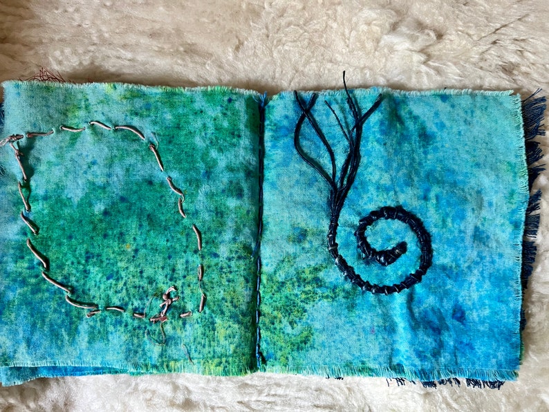 Puede incluir: Un diario de tela hecho a mano con una cubierta de m&aacute;rmol azul y verde. El diario tiene un c&iacute;rculo cosido en la p&aacute;gina izquierda y una espiral de hilo negro en la p&aacute;gina derecha.