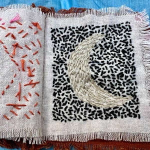 Puede incluir: Un cuadrado de tela blanca con un dise&ntilde;o de luna creciente bordado en blanco y negro. La tela tiene un borde deshilachado.