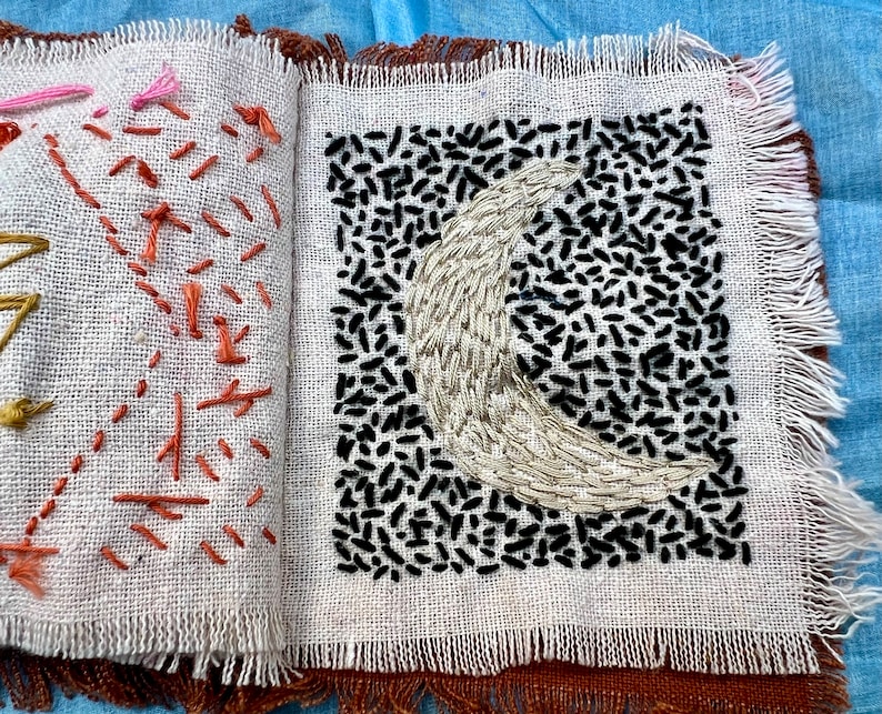 Puede incluir: Un cuadrado de tela blanca con un dise&ntilde;o de luna creciente bordado en blanco y negro. La tela tiene un borde deshilachado.