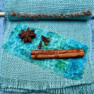Puede incluir: Una tela de arpillera azul con un borde cosido marr&oacute;n. Un trozo de tela azul con tinte verde y azul est&aacute; encima de la arpillera. Sobre la tela hay dos palitos de canela, una estrella de an&iacute;s y cuatro clavos.