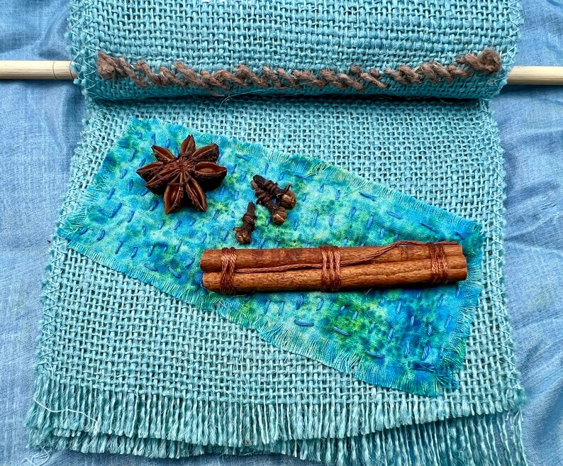 Puede incluir: Una tela de arpillera azul con un borde cosido marr&oacute;n. Un trozo de tela azul con tinte verde y azul est&aacute; encima de la arpillera. Sobre la tela hay dos palitos de canela, una estrella de an&iacute;s y cuatro clavos.