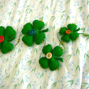以下が含まれることがあります： ビーズでつなげられた、ボタンの中心を持つ緑色のフェルト製のクローバーの形が4つ。ボタンは赤、青、オレンジ、白です。