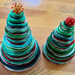 May include: Two felt Christmas trees, one larger than the other, are made of stacked circles of red, green, and white felt. The tops of the trees are decorated with red and gold beads.