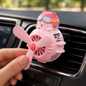 May include: A pink car air freshener vent clip featuring a cartoon character wearing goggles and a helmet, with a rotating propeller fan on the front. The body is printed with the text SPACE DCABLE FAMILY and the number #01 #01 in black ink.