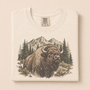 May include: A cream-colored t-shirt featuring a graphic print of a brown bison standing in front of a forest of pine trees and mountain peaks. The shirt has a Comfort Colors brand label at the neckline with the size M printed on the tag.