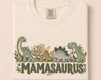Könnte beinhalten: Ein cremefarbenes T-Shirt mit einer Grafik von vier grünen und grauen Dinosauriern über dem Schriftzug MAMASAURUS in dunkelgrünen Buchstaben. Das T-Shirt hat ein Comfort Colors Markenetikett am Kragen, das die Größe M angibt, und zeigt Blattmotive.