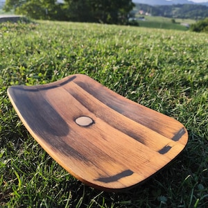 May include: A rectangular wooden tray with curved edges, featuring a natural wood grain pattern with alternating light tan and dark brown streaks. A small, circular, light-coloured wooden inlay is centred on the surface of the tray.