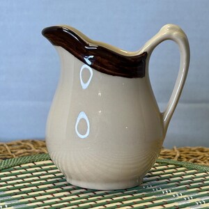May include: A small ceramic creamer pitcher with a glossy beige finish and a dark brown painted rim. The pitcher features a curved handle on the right side and a distinct pouring spout. The surface is smooth with a reflective glaze throughout.