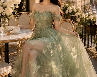 May include: A floor-length sage green off-the-shoulder dress featuring a sheer tulle overlay adorned with intricate embroidered butterfly appliques, a sweetheart neckline, and a flowing skirt with a high leg slit, crafted from layered fabric.