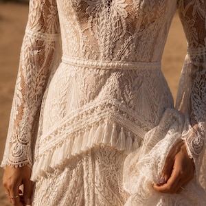 May include: A long-sleeved ivory lace dress featuring a deep V-neckline, intricate floral patterns, and a fitted waistline accented with a horizontal band. The skirt includes a layered section adorned with a row of delicate hanging fringe tassels.