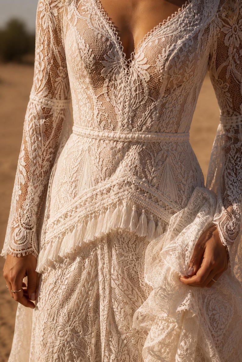 May include: A long-sleeved ivory lace dress featuring a deep V-neckline, intricate floral patterns, and a fitted waistline accented with a horizontal band. The skirt includes a layered section adorned with a row of delicate hanging fringe tassels.