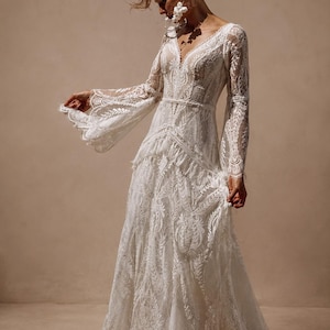 May include: A floor-length white lace dress featuring long, flared sleeves, a deep V-neckline, and a fitted bodice with a defined waistline. The garment is constructed from intricate floral lace patterns throughout, with a flowing skirt.