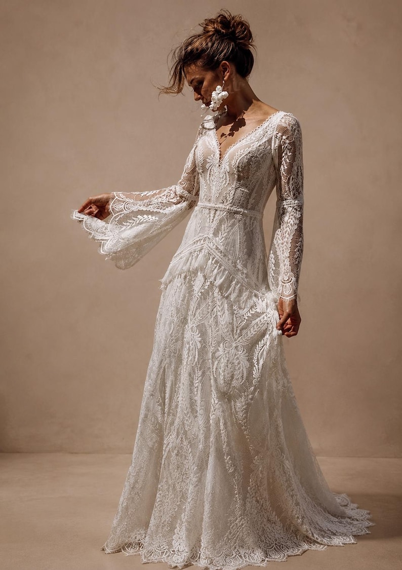 May include: A floor-length white lace dress featuring long, flared sleeves, a deep V-neckline, and a fitted bodice with a defined waistline. The garment is constructed from intricate floral lace patterns throughout, with a flowing skirt.