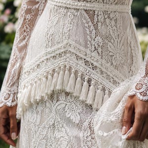 May include: A long-sleeved ivory lace dress featuring an intricate floral pattern, a fitted waistline with a horizontal band, and a V-shaped overlay panel adorned with a row of hanging fringe tassels along the bottom edge of the bodice.