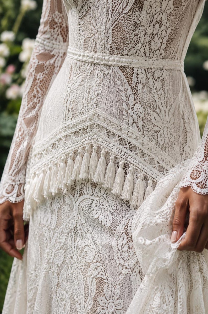 May include: A long-sleeved ivory lace dress featuring an intricate floral pattern, a fitted waistline with a horizontal band, and a V-shaped overlay panel adorned with a row of hanging fringe tassels along the bottom edge of the bodice.