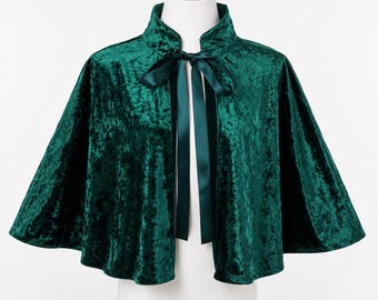 May include: A dark emerald green velvet capelet featuring a high collar and a matching satin ribbon tie closure at the neckline. The fabric has a crushed texture that catches the light, and the garment drapes in a wide, flared silhouette.