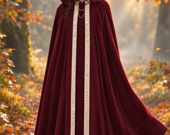 May include: A floor-length deep burgundy hooded cloak featuring a wide, cream-colored decorative trim along the front opening and hood edge. The garment is secured at the neck with a gold-toned metal clasp and a delicate double-chain detail.