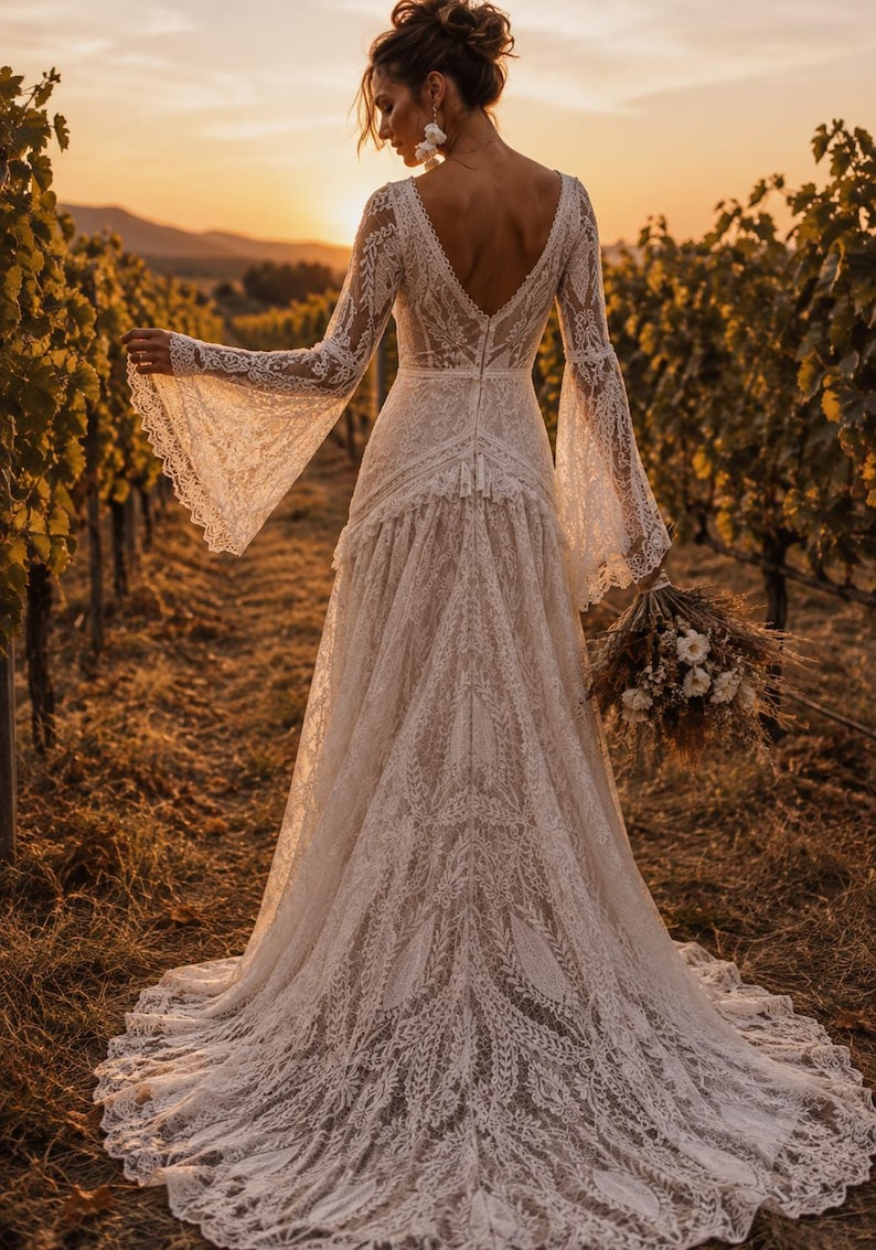 May include: A floor-length white lace dress featuring long bell sleeves, a deep V-neckline, and a fitted bodice that transitions into a flowing skirt with a train. The intricate lace pattern covers the entire garment, including the flared sleeves.