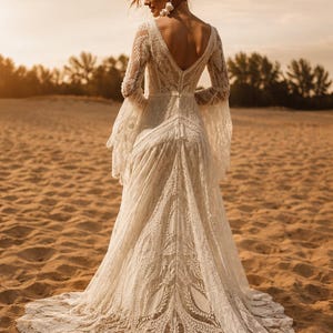 May include: A floor-length off-white lace dress featuring long bell sleeves, a deep V-neckline on the back, and an intricate floral pattern throughout the fabric. The garment has a fitted bodice and a flowing skirt that extends into a train.