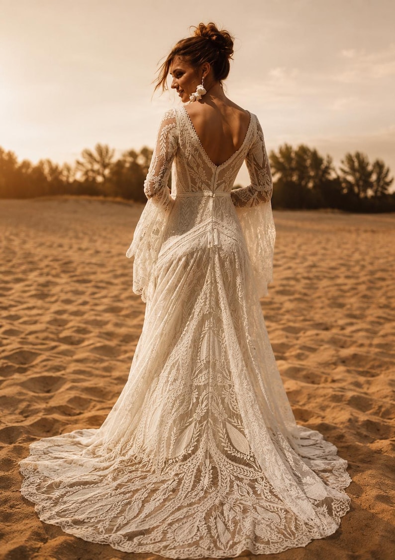 May include: A floor-length off-white lace dress featuring long bell sleeves, a deep V-neckline on the back, and an intricate floral pattern throughout the fabric. The garment has a fitted bodice and a flowing skirt that extends into a train.