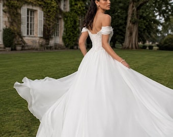 May include: A white off-the-shoulder wedding dress with a sweetheart neckline and a corset-style bodice. The gown features a voluminous, floor-length skirt made of layered, flowing fabric that flares out, creating a wide, circular silhouette.