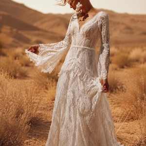 May include: A floor-length white lace dress featuring long bell sleeves, a V-neckline, and a fitted bodice with a layered skirt. The intricate floral lace pattern covers the entire garment, which includes a defined waistline and flared cuffs.