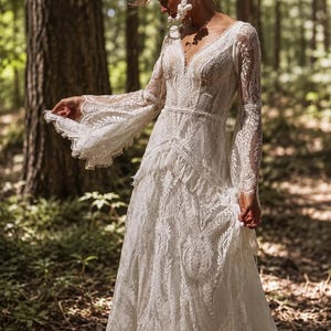 May include: A floor-length white lace gown featuring long, flared bell sleeves, a deep V-neckline, and a fitted bodice with a defined waistline. The dress is crafted from intricate floral lace patterns with scalloped edges and a tiered skirt.
