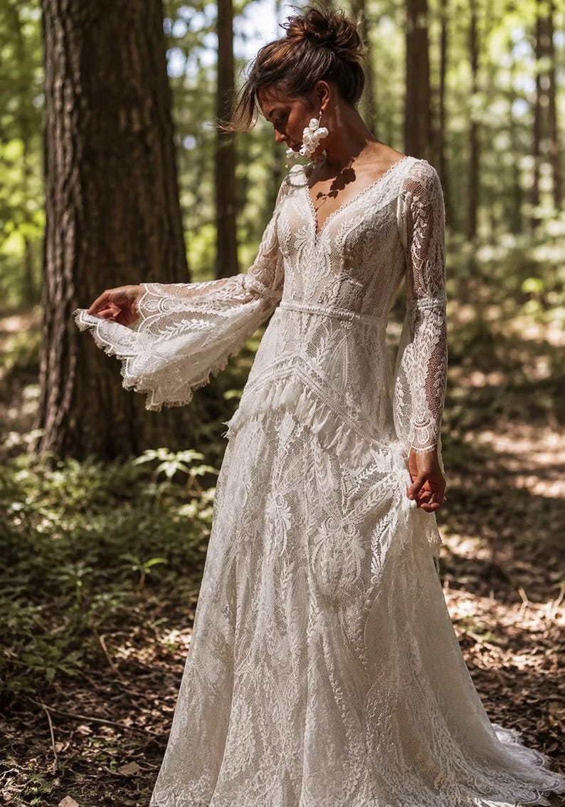 May include: A floor-length white lace gown featuring long, flared bell sleeves, a deep V-neckline, and a fitted bodice with a defined waistline. The dress is crafted from intricate floral lace patterns with scalloped edges and a tiered skirt.