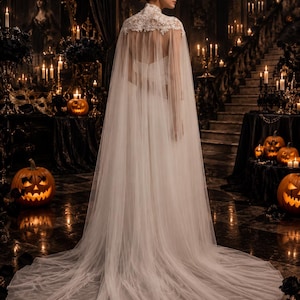 May include: A floor-length white bridal cape featuring a high neck with intricate lace detailing at the shoulders and upper back. The garment is crafted from sheer, flowing tulle fabric that extends into a long, sweeping train behind the wearer.