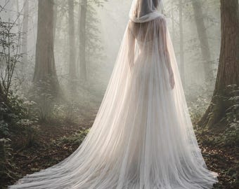 May include: A floor-length white bridal gown featuring a sheer, flowing tulle cape that extends into a long train. The garment includes a hood made of the same lightweight, translucent fabric, draping elegantly over the shoulders and back.