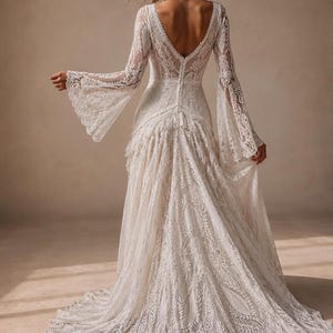 May include: A floor-length white lace dress featuring long bell sleeves, a deep V-shaped back neckline, and a row of small buttons running down the center back. The fabric displays an intricate floral pattern throughout, ending in a full train.