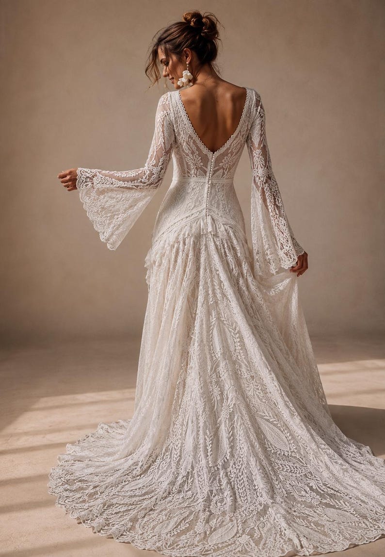 May include: A floor-length white lace dress featuring long bell sleeves, a deep V-shaped back neckline, and a row of small buttons running down the center back. The fabric displays an intricate floral pattern throughout, ending in a full train.